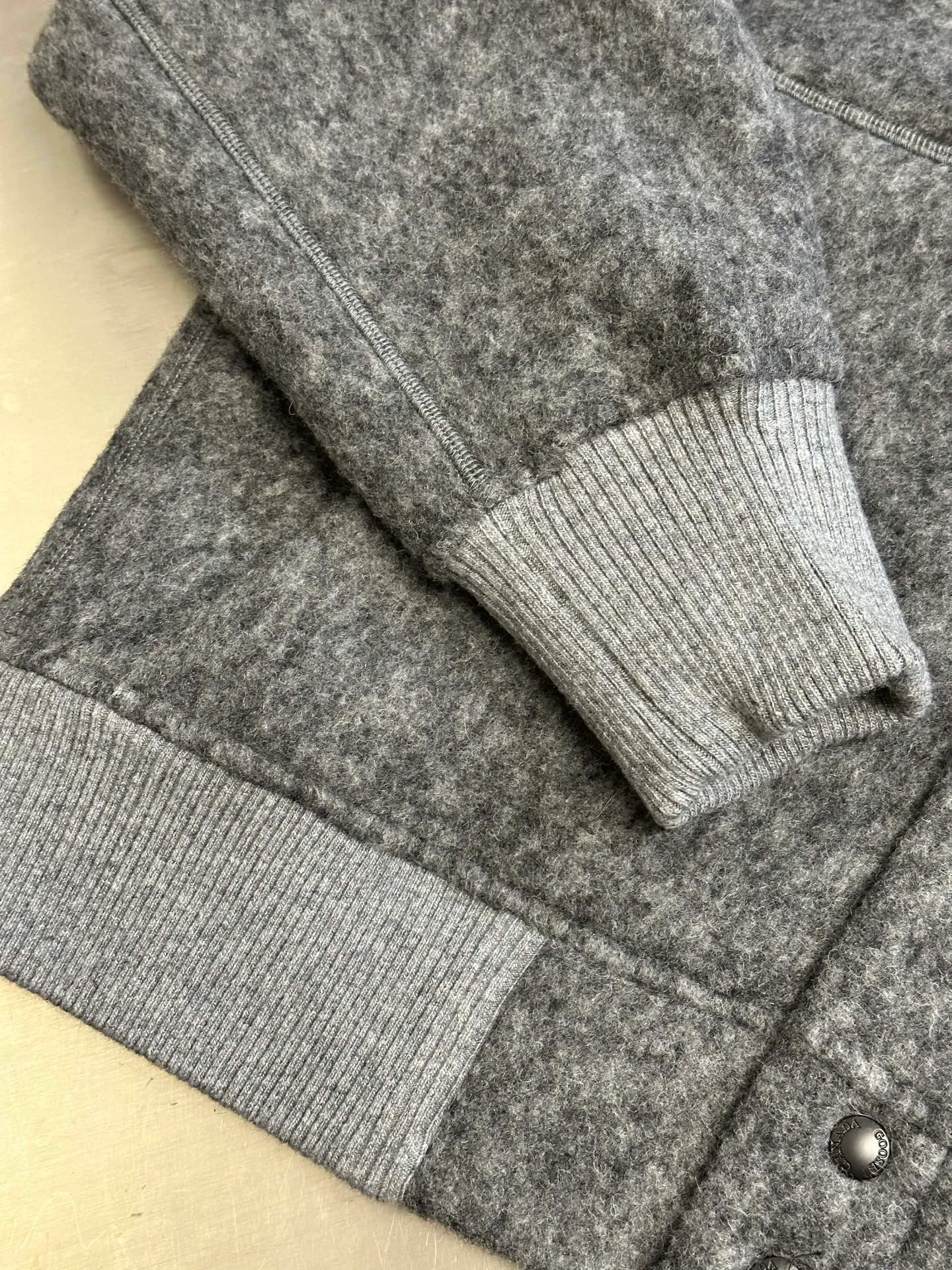 P770 Canada Goose Fleece Badge Button Jacket The main fabric of this jacket is light gray wool blend fabric to improve the fabric's wear resistance, wrinkle resistance and warmth. Wool provides good warmth and a natural feel, while blended fibers enhance the fabric's practicality and durability.  The cutting process needs to ensure that the texture and color of the fabric are evenly distributed, especially with this light gray tone, to avoid chromatic aberrations. The stretchability of wool-blend fabrics needs to be taken into consideration when cutting pieces to ensure dimensional stability of the finished garment.  Use a sewing needle and thread suitable for wool-blend fabrics to avoid damage to the fabric during sewing. The sewing stitches are even and firm to prevent thread stripping.  The temperature needs to be controlled during ironing to avoid high temperature damage to the fabric while keeping the fabric smooth and crisp. The front and back body parts, sleeves and other main parts are stitched with flat seams to ensure a smooth silhouette. Align the fabric texture at the splices to keep the look neat.  The white circular armband on the left arm is an important part of the brand identity and is finely sewn to ensure it is strong and flat.  size：s m l xl